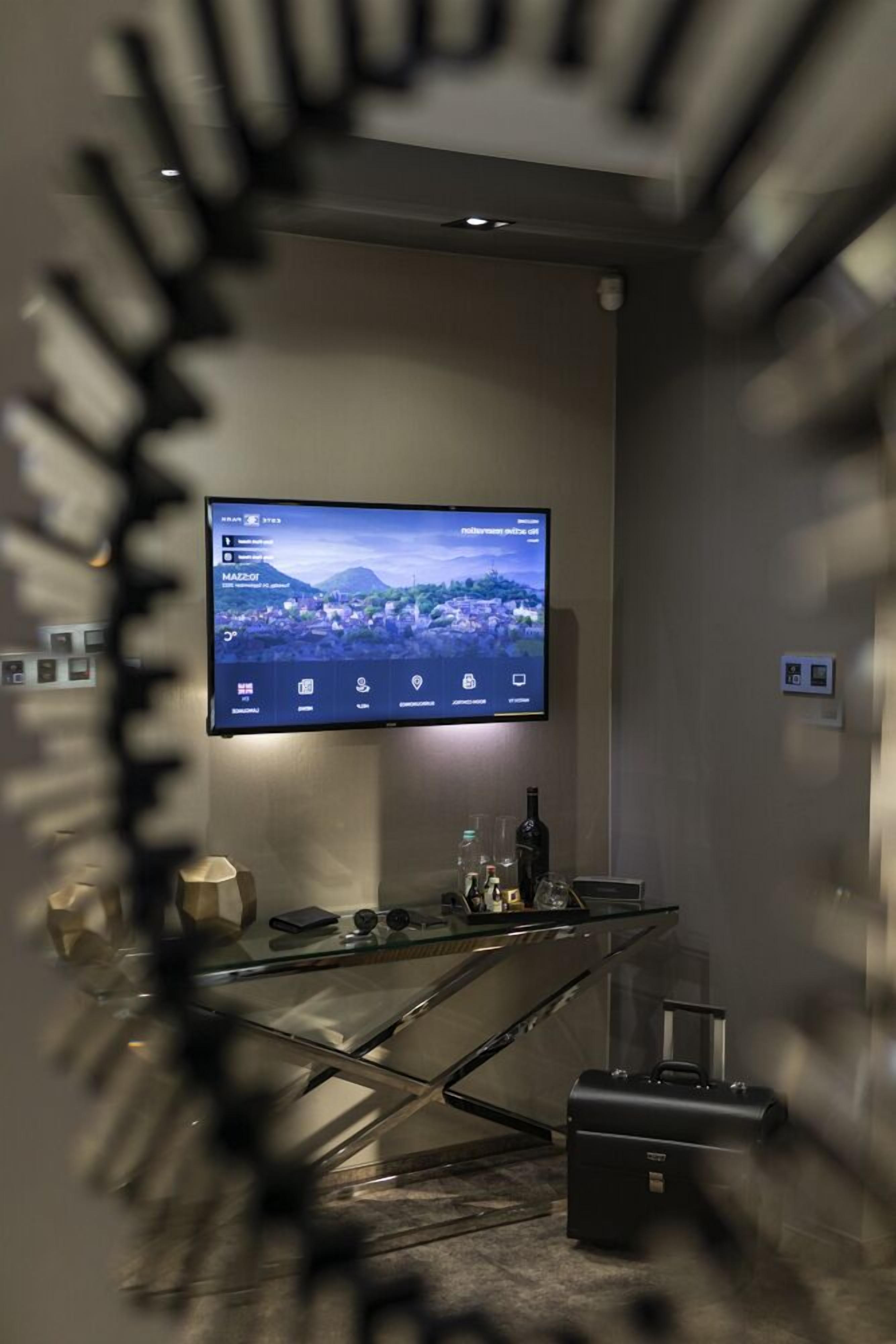 A modern hotel room with a large flat-screen TV mounted on the wall, a glass desk with a laptop, a bottle, and a suitcase.