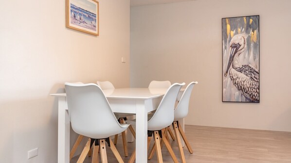 Un coin repas avec une table et des chaises blanches, un tableau encadré représentant des oiseaux et un mur de couleur claire.