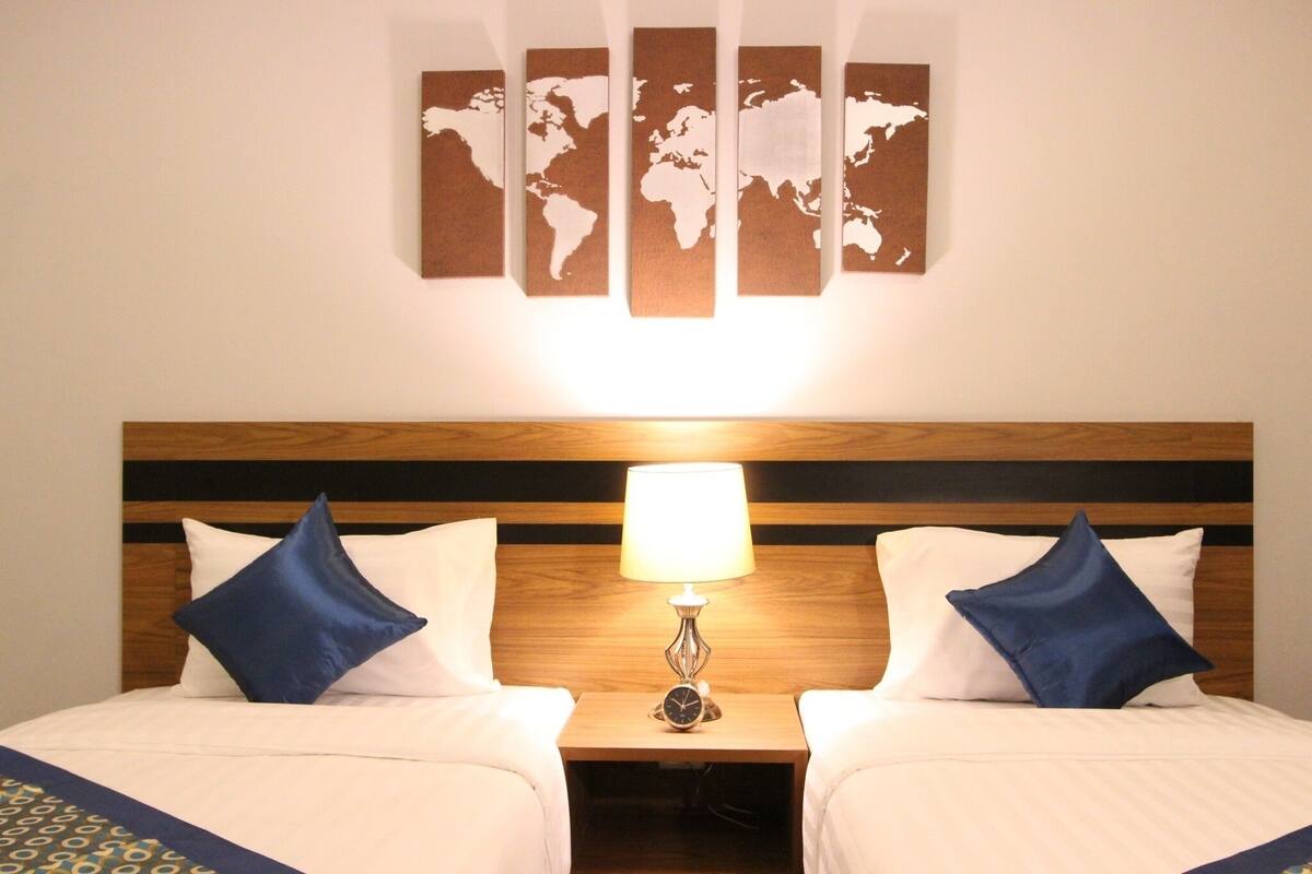 Una habitación de hotel con cabecera de madera, dos camas con almohadas azules, una mesita de noche con lámpara y un mapa del mundo en la pared.