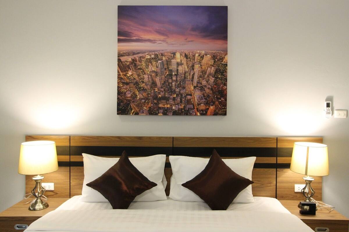 Habitación de hotel con vista a la ciudad, cabecera de madera, dos lámparas de noche y dos almohadas marrones.