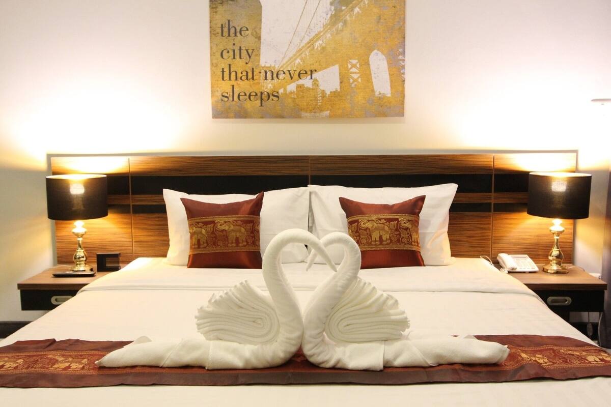 Habitación de hotel con cama, decoración de toallas en forma de cisne, cabecera de madera y dos mesitas de noche con lámparas.