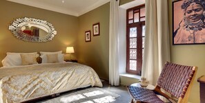 Een slaapkamer met een groot bed, een decoratieve spiegel, een raam met gordijnen, en een ingelijst schilderij aan de muur.