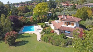 A large house with a swimming pool, surrounded by lush greenery and other houses in the background.