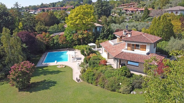 A large house with a swimming pool, surrounded by lush greenery and other houses in the background. - Villa Gelso (Soiano del Lago)