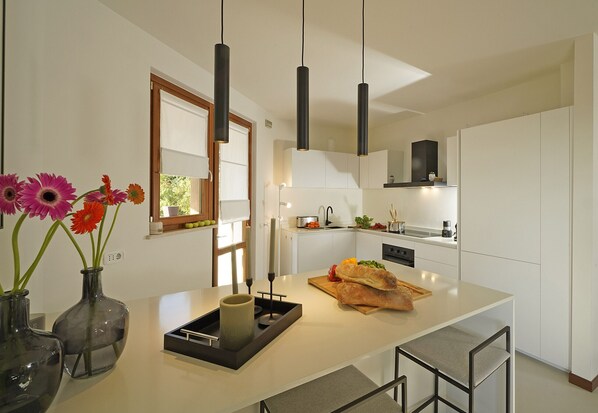 A modern kitchen with white cabinets, a central island, pendant lights, and a window with a view of greenery. - Villa Gelso (Soiano del Lago)