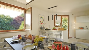 A dining area with a table set for a meal, a kitchen in the background, and a view of greenery outside.