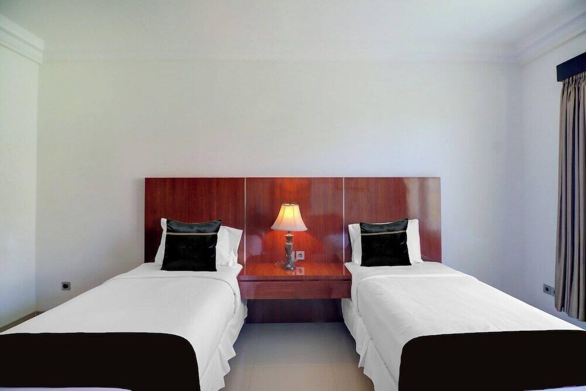 Ein Hotelzimmer mit zwei Einzelbetten, einem Holzkopfstück, einem Nachttisch mit Lampe und einem Fenster mit Vorhängen.