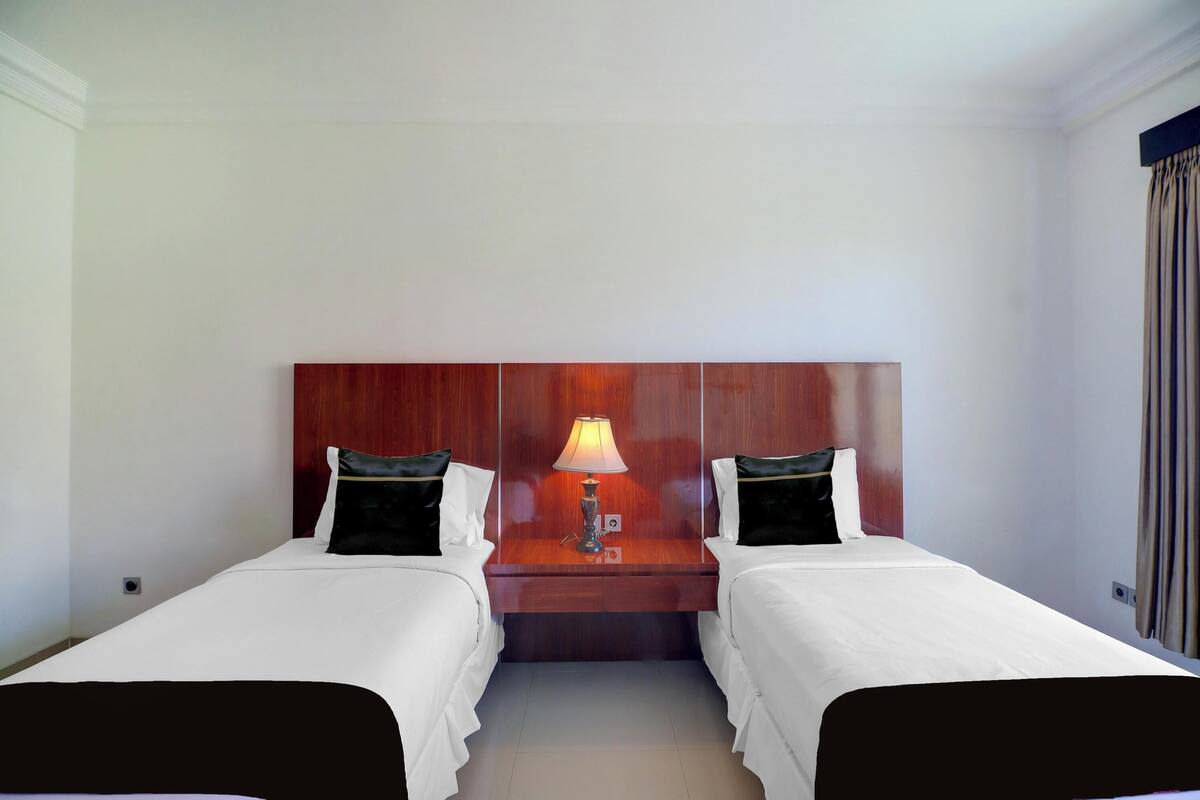 Ein Hotelzimmer mit zwei Einzelbetten, einem Holzkopfstück, einem Nachttisch mit Lampe und einem Fenster mit Vorhängen.