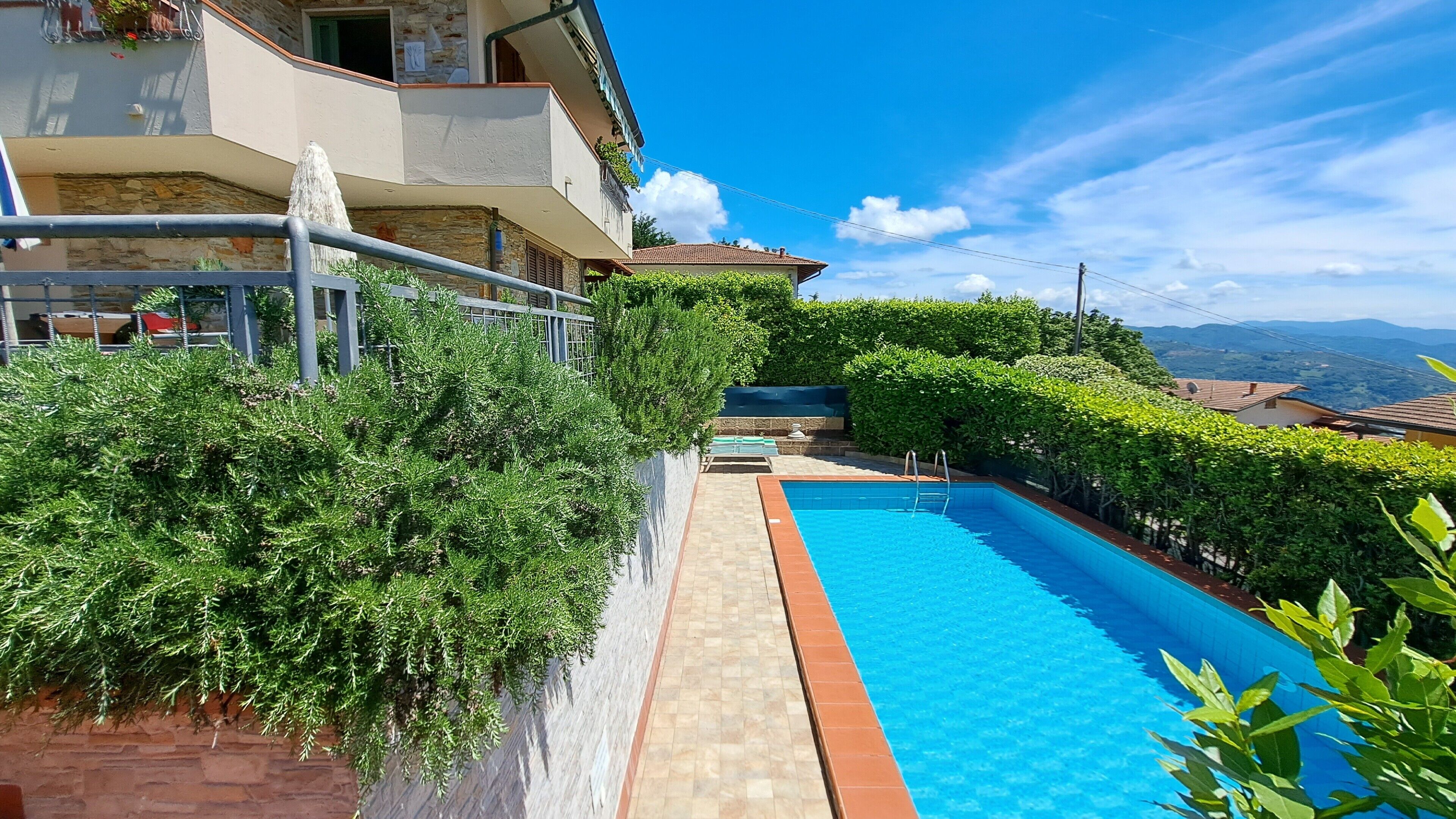 A swimming pool with a tiled deck, surrounded by lush greenery and a stone wall.