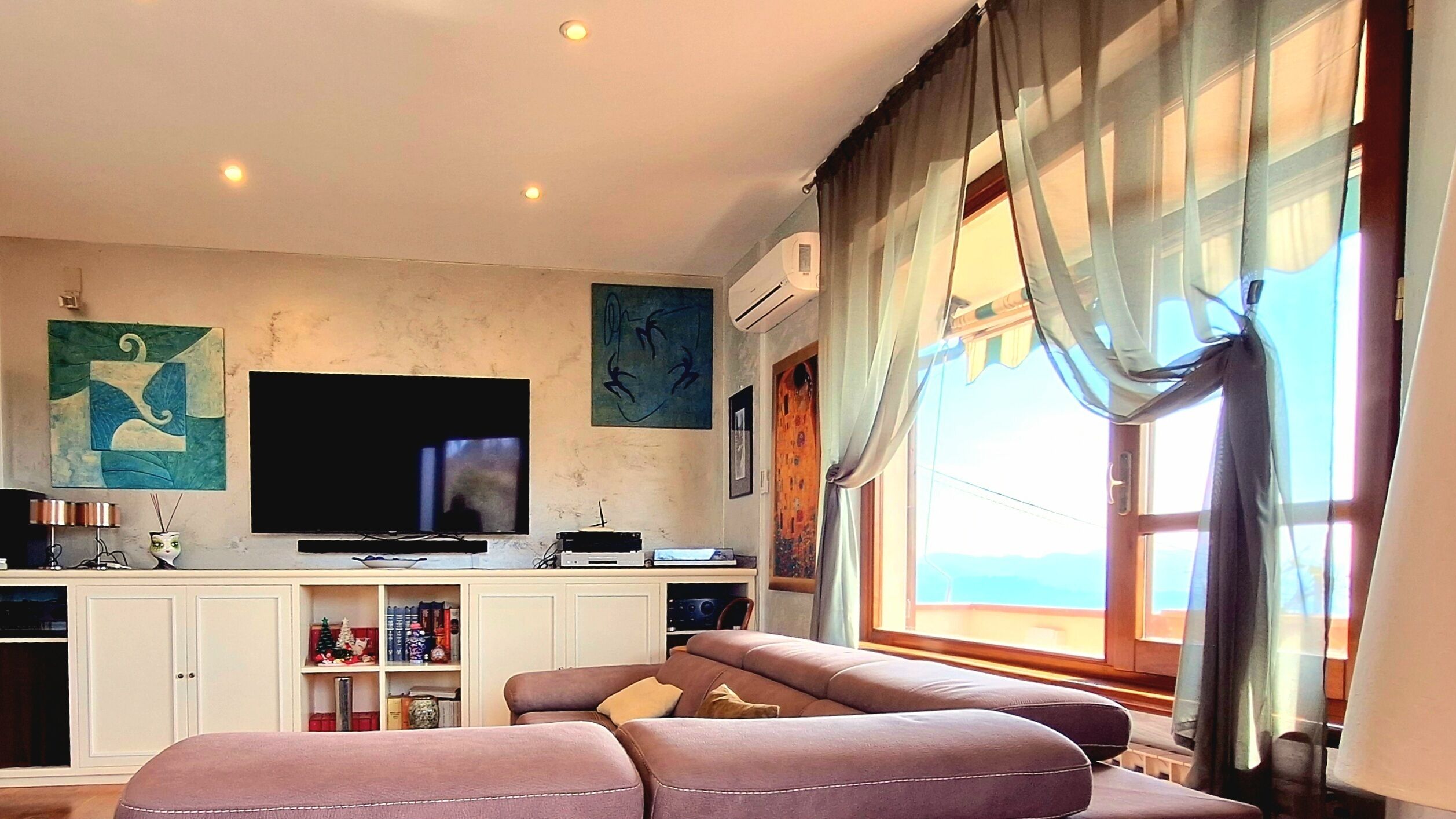A living room with a flat-screen TV, a sofa, a cabinet with books, and a window with curtains.
