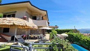 Een twee verdiepingen tellend huis met een stenen gevel, een balkon met een rieten zonnescherm, zitplaatsen buiten en een zwembad.