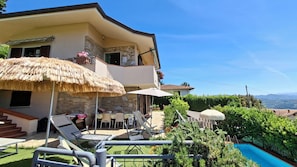 A two-story house with a stone facade, a balcony with a thatched umbrella, outdoor seating, and a swimming pool.