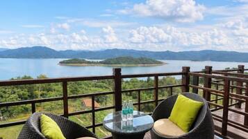 A balcony with wicker chairs and a table, overlooking a lake and mountains.