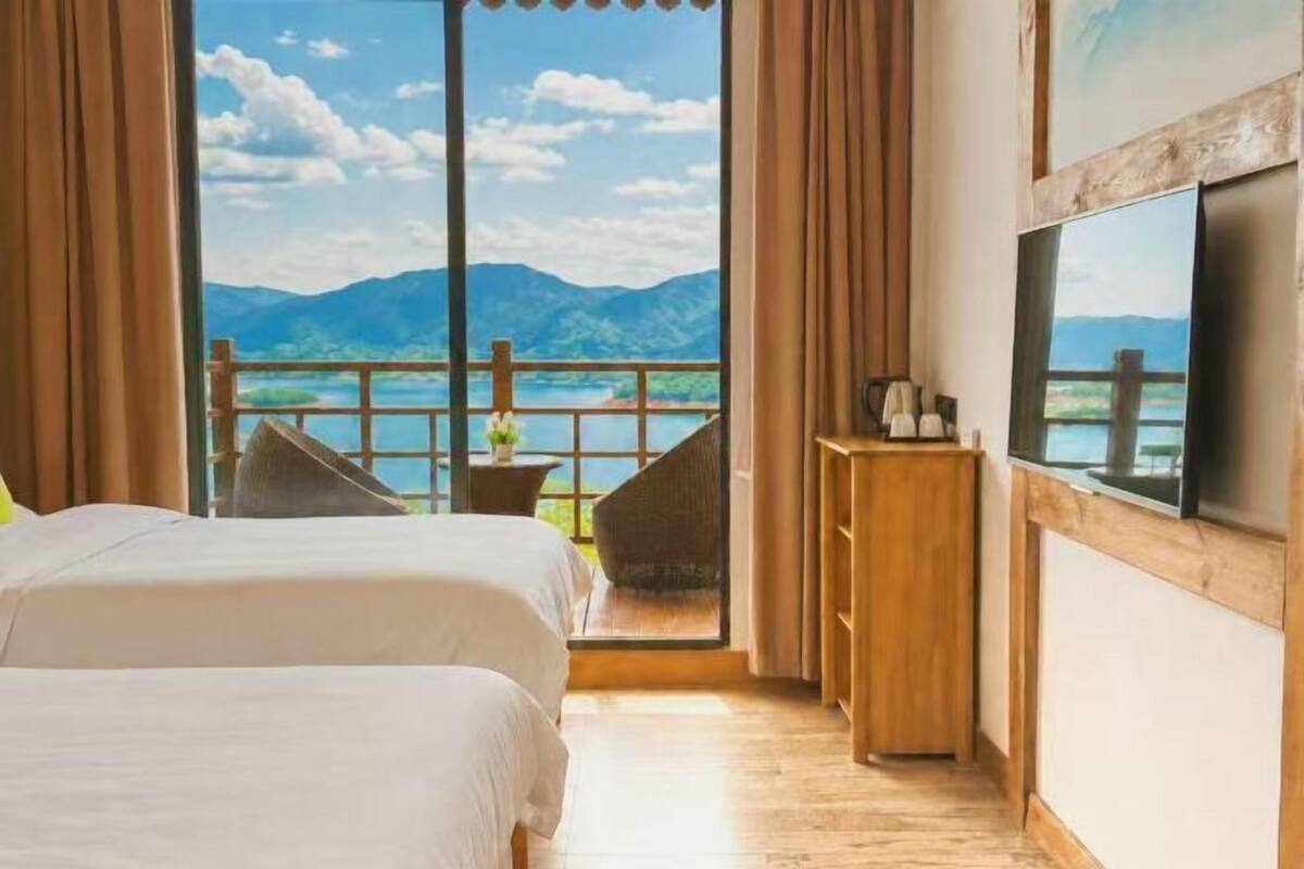 A bedroom with two beds, a wooden dresser, a TV, and a balcony with a view of mountains and water.