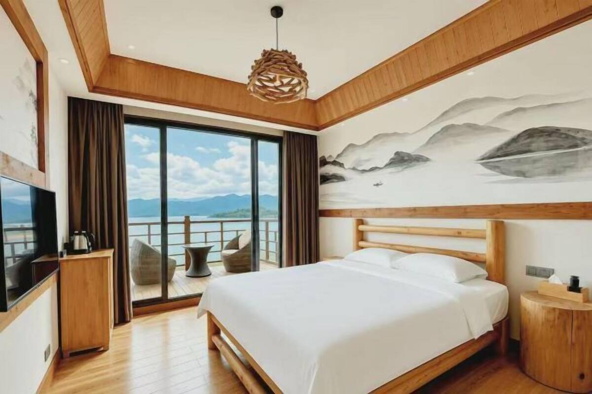 A bedroom with a large bed, wooden ceiling, a painting of mountains, a TV, and a view of the sea.