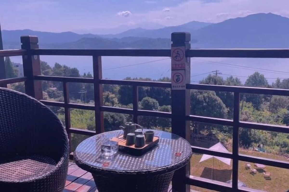 A balcony with wicker furniture, a table with teapots and cups, and a view of mountains and greenery.