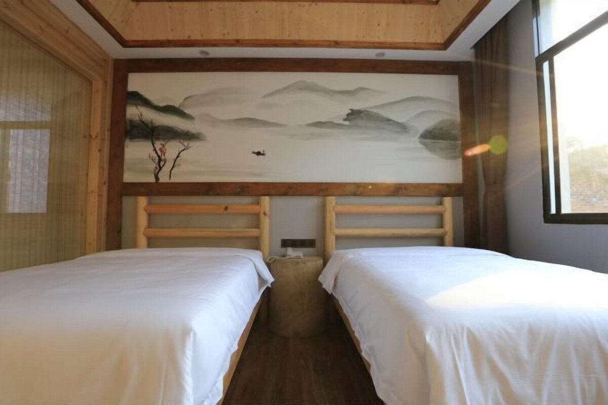 A room with two beds, wooden headboards, a large mural of mountains, and a window on the right.