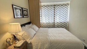 A neatly made bed with white bedding, a bedside lamp, and a wooden nightstand with a phone and charger. - Enjoy Lovely 4 bed Apartment With Garden (London)