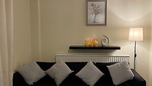 A black sofa with white cushions, a radiator, a shelf with candles, and a framed picture on the wall. - Enjoy Lovely 4 bed Apartment With Garden (London)