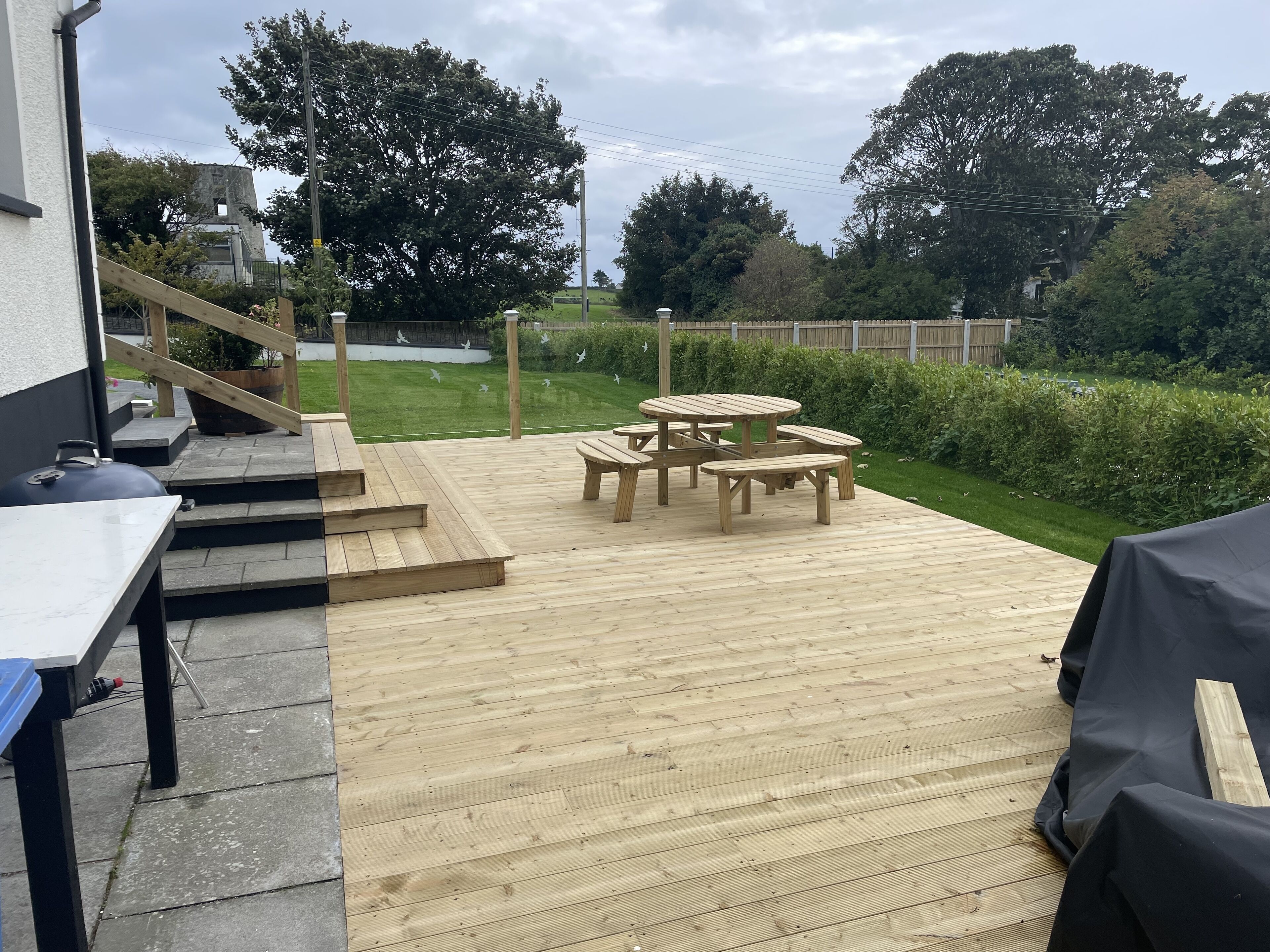 Une terrasse en bois avec des meubles d’extérieur, un barbecue et une vue sur une cour verdoyante bordée d’arbres.