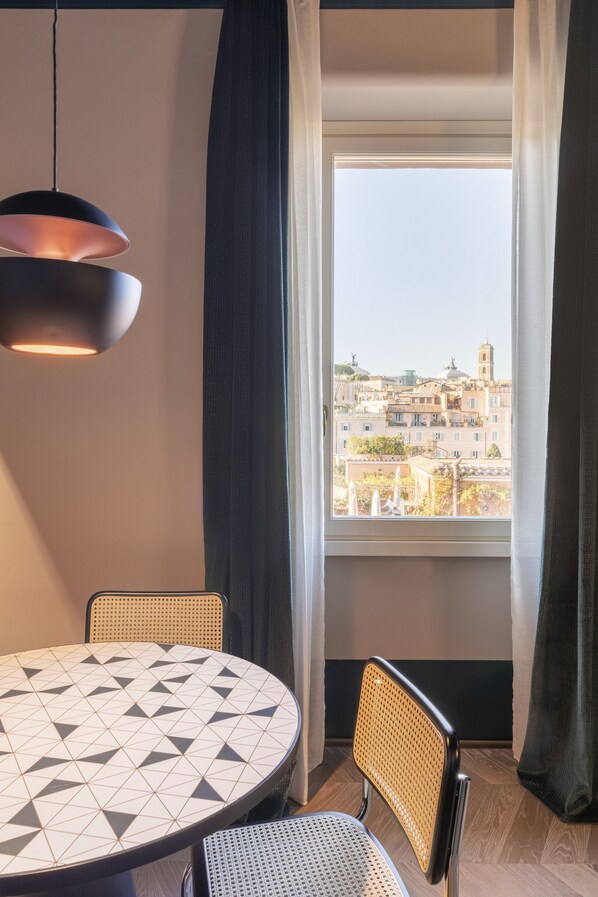 Quarto com uma mesa redonda, duas cadeiras, luminária pendente e vista para o horizonte da cidade através de uma janela com cortinas.