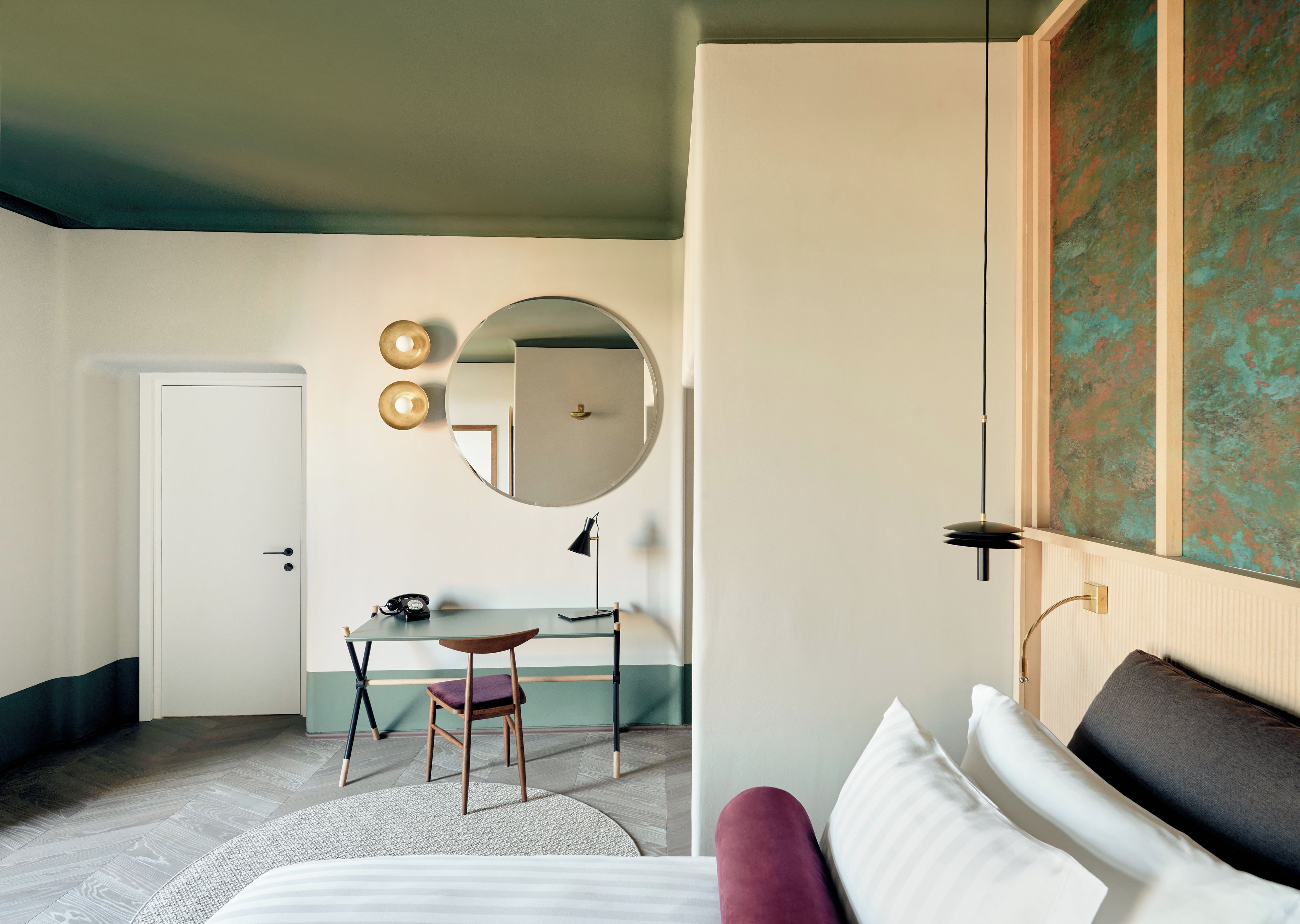 Quarto de hotel com uma cama, uma escrivaninha com cadeira, um espelho redondo e uma porta.