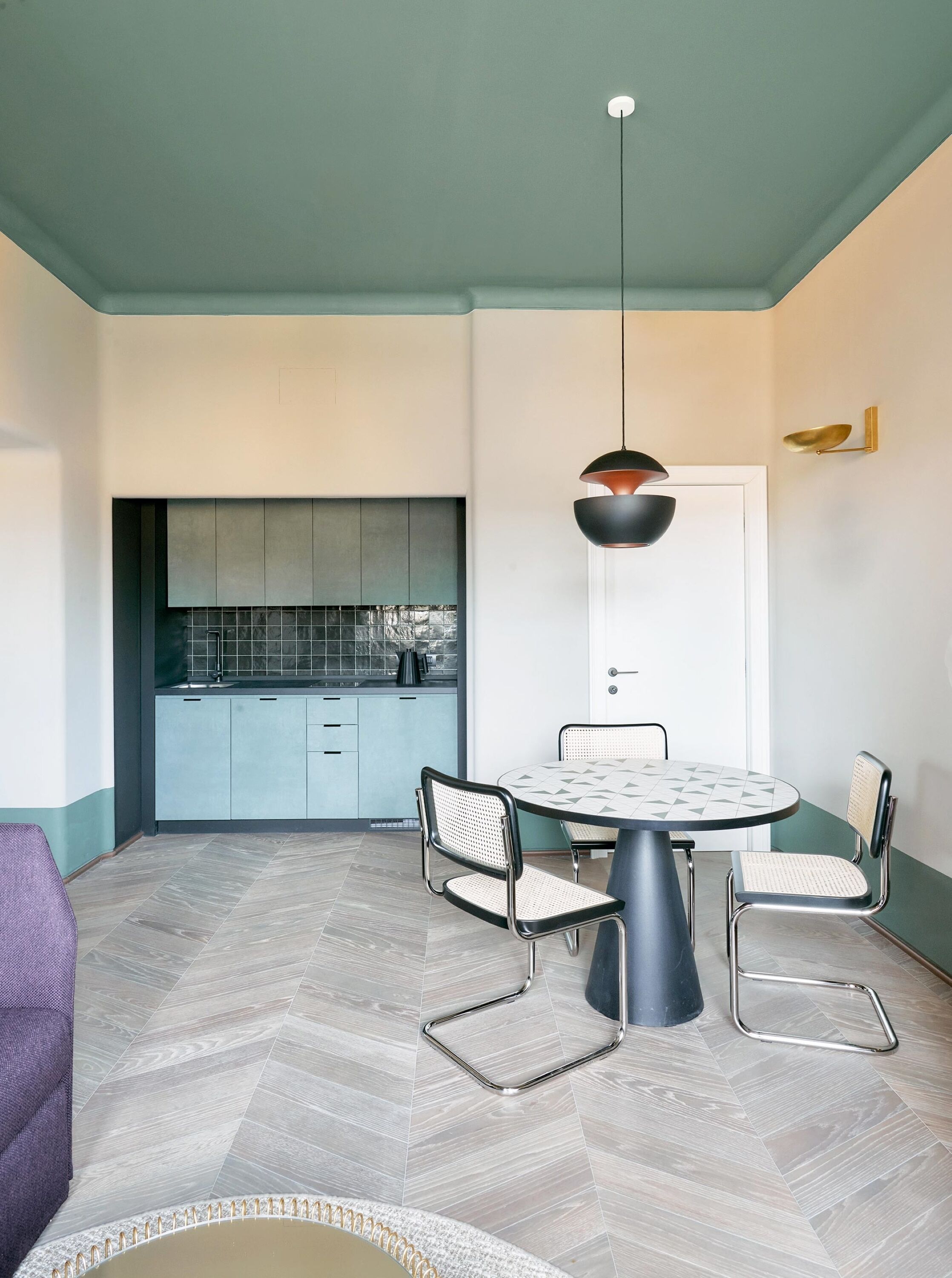 A modern kitchen with light blue cabinets and a white door. There is a round dining table with a geometric tile top, surrounded by metal chairs. A pendant light hangs above the table.