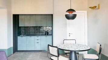 A modern kitchen with light blue cabinets and a white door. There is a round dining table with a geometric tile top, surrounded by metal chairs. A pendant light hangs above the table.