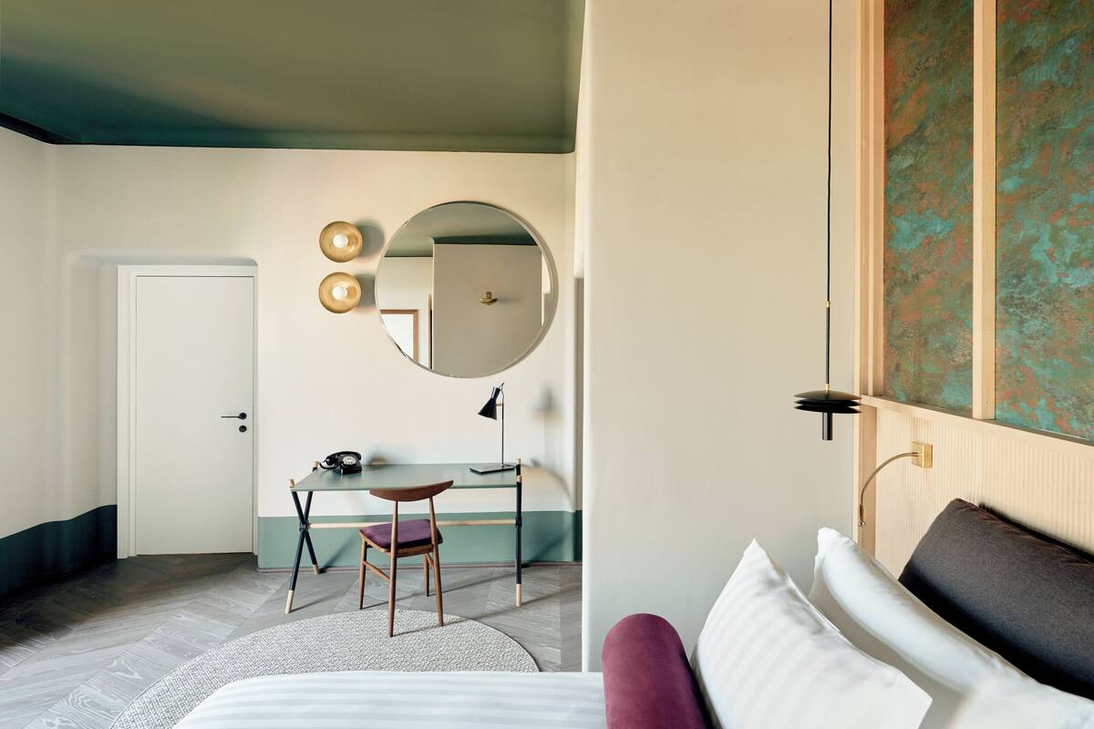 A hotel room with a bed, a desk with a chair, a round mirror, and a door.