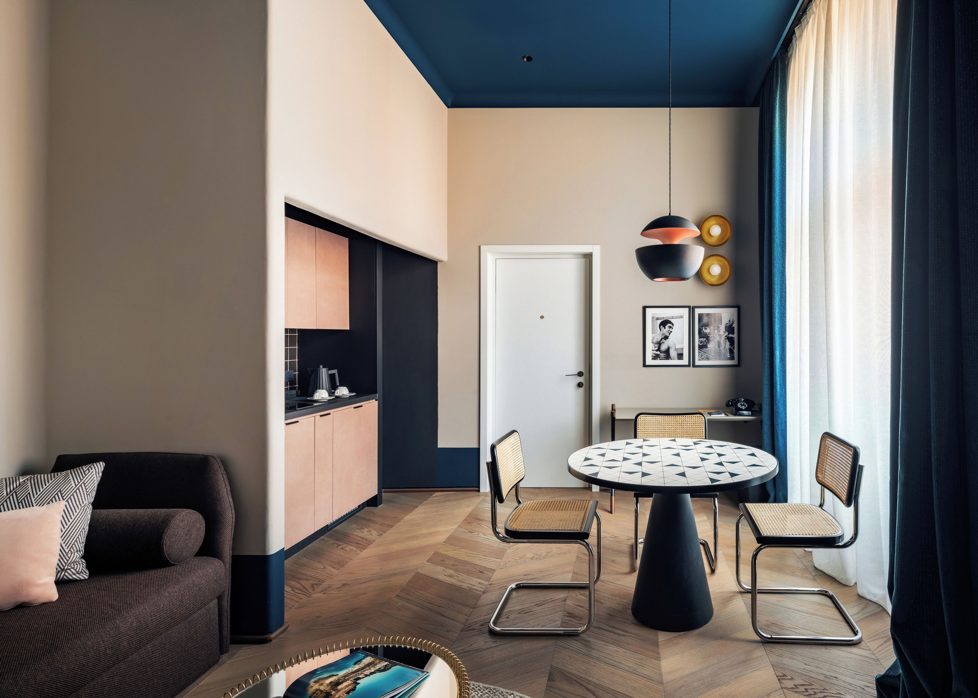 A modern living room with a sofa, a dining table with chairs, a kitchen area, and a large window with curtains.