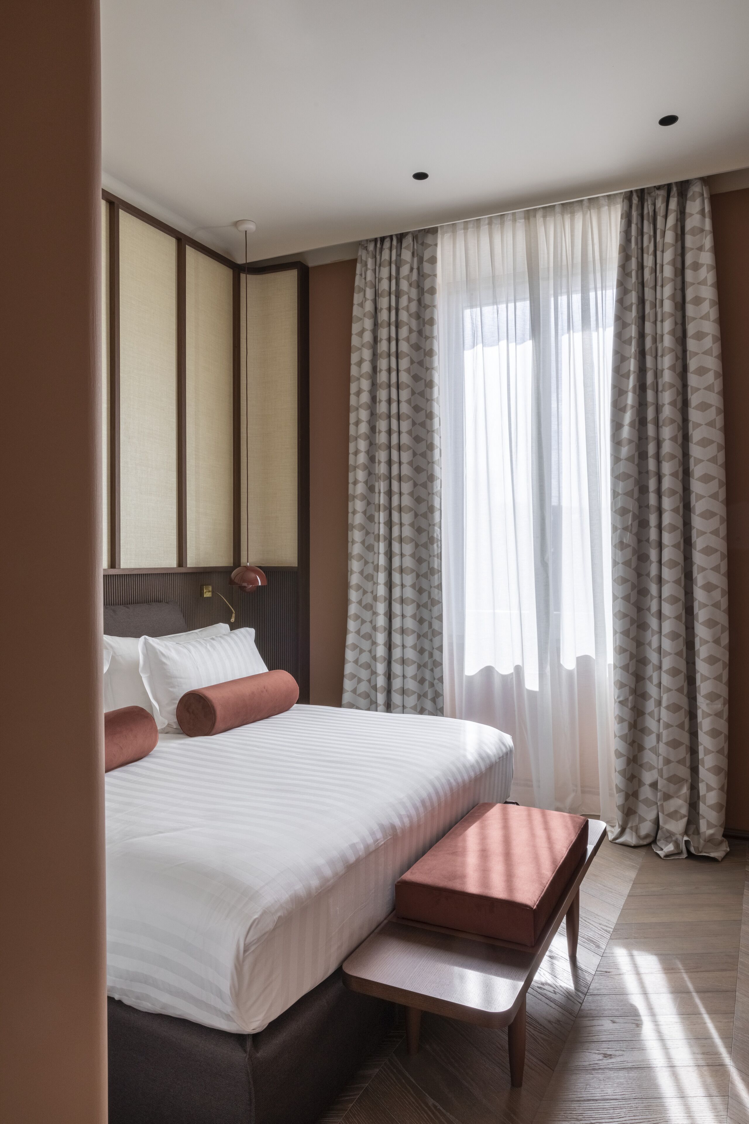 A hotel room with a large bed, a bedside table, a bench, a lamp, and a window with curtains.
