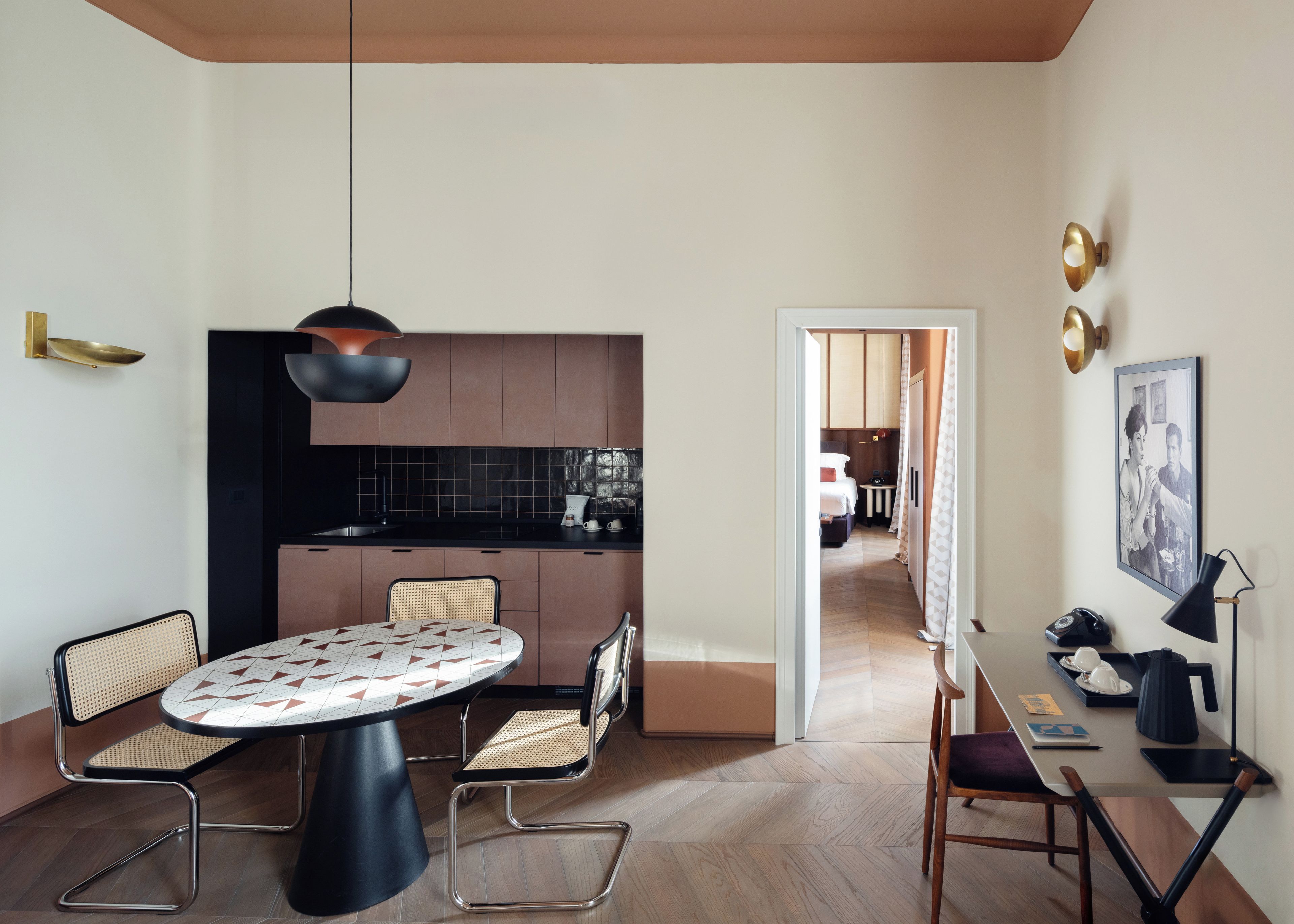 A modern interior with a dining area featuring a round table and chairs, a kitchen with dark cabinetry, and a hallway leading to a bedroom.
