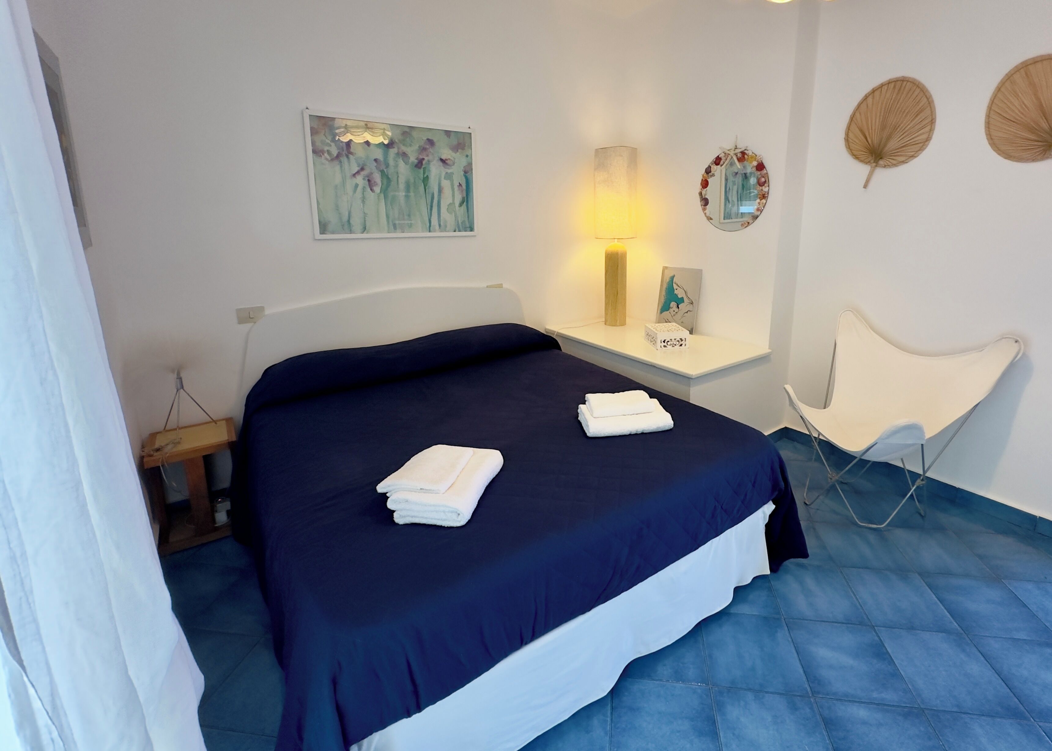 Un lit bien fait, recouvert d’une couvre-lit bleu foncé, et deux serviettes blanches pliées sont posées dessus. On aperçoit une table de chevet avec une lampe et un objet décoratif.