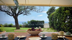 Tavola imbandita con una ciotola di frutta, piatti e posate, con vista su un panorama che include mare e giardino.