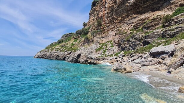Paesaggio costiero con acque turchesi cristalline, scogliere rocciose e una spiaggia sabbiosa.