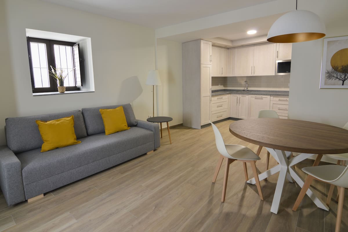 Un salon moderne avec un canapé gris, des coussins jaunes, une table de salle à manger ronde en bois et des chaises blanches.