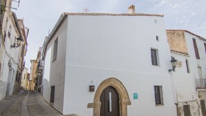 A narrow street with a white building featuring a dark arched entrance, a small balcony, and a sign indicating an address. - Apartamentos Turísticos Hornillo (Cáceres)