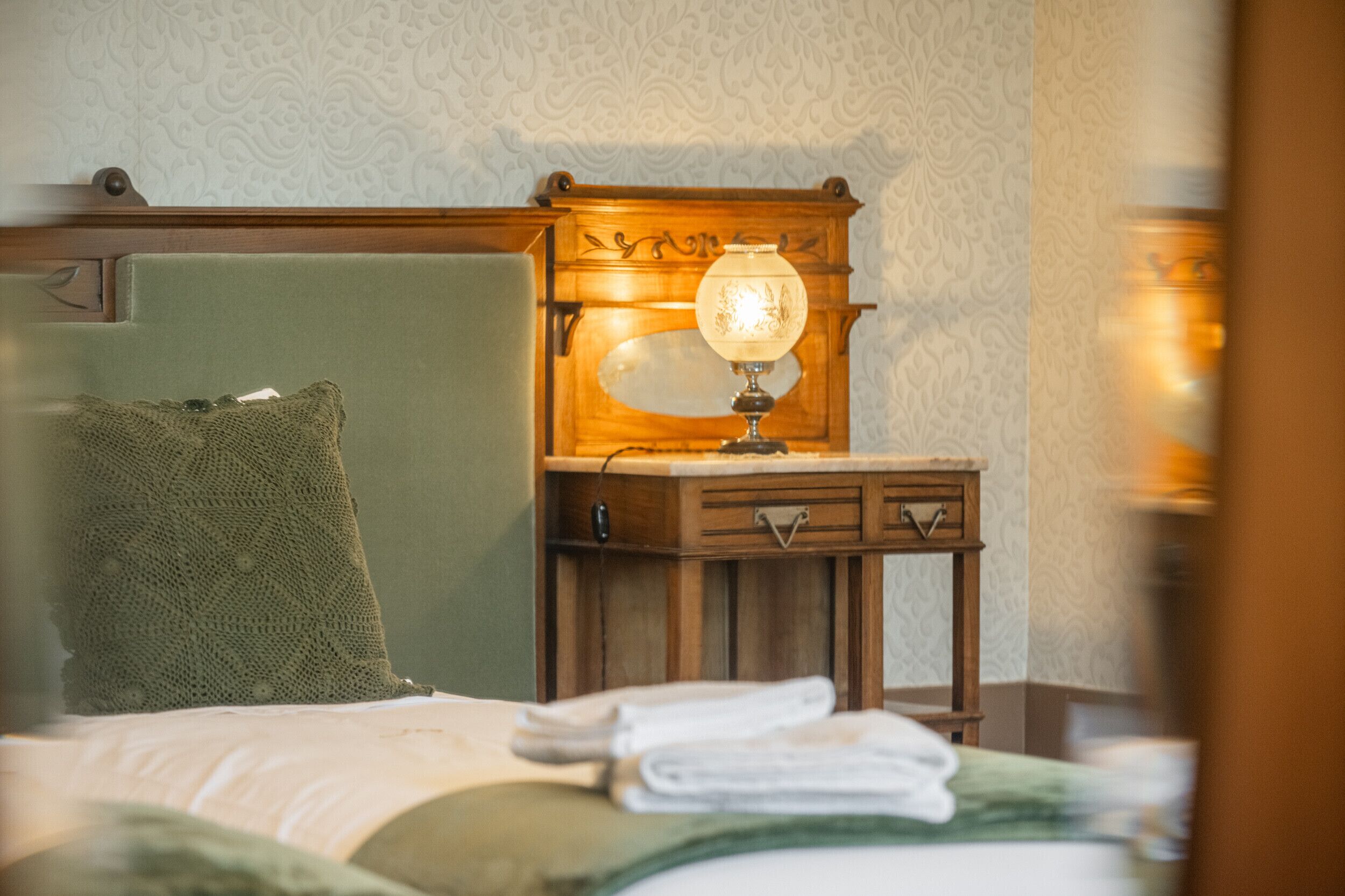 A neatly made bed with a green pillow, a wooden nightstand with a lamp, and a wall with a patterned wallpaper.