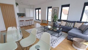 A modern living room with a grey sofa, a glass coffee table, and white chairs. There is a kitchen area with white cabinets and a dining table with chairs.