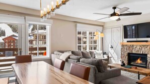 A modern living room with a fireplace, a large window with a view of a snowy mountain, and a dining area with a wooden table and chairs. - RARE Three Bedroom Lovely Passage Point Condo!! (Copper Mountain)