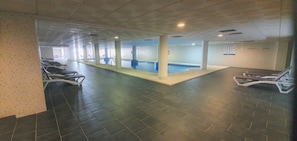 Un vaste espace intérieur abritant une piscine avec plusieurs couloirs de nage, un plongeoir et un coin salon équipé de chaises.
