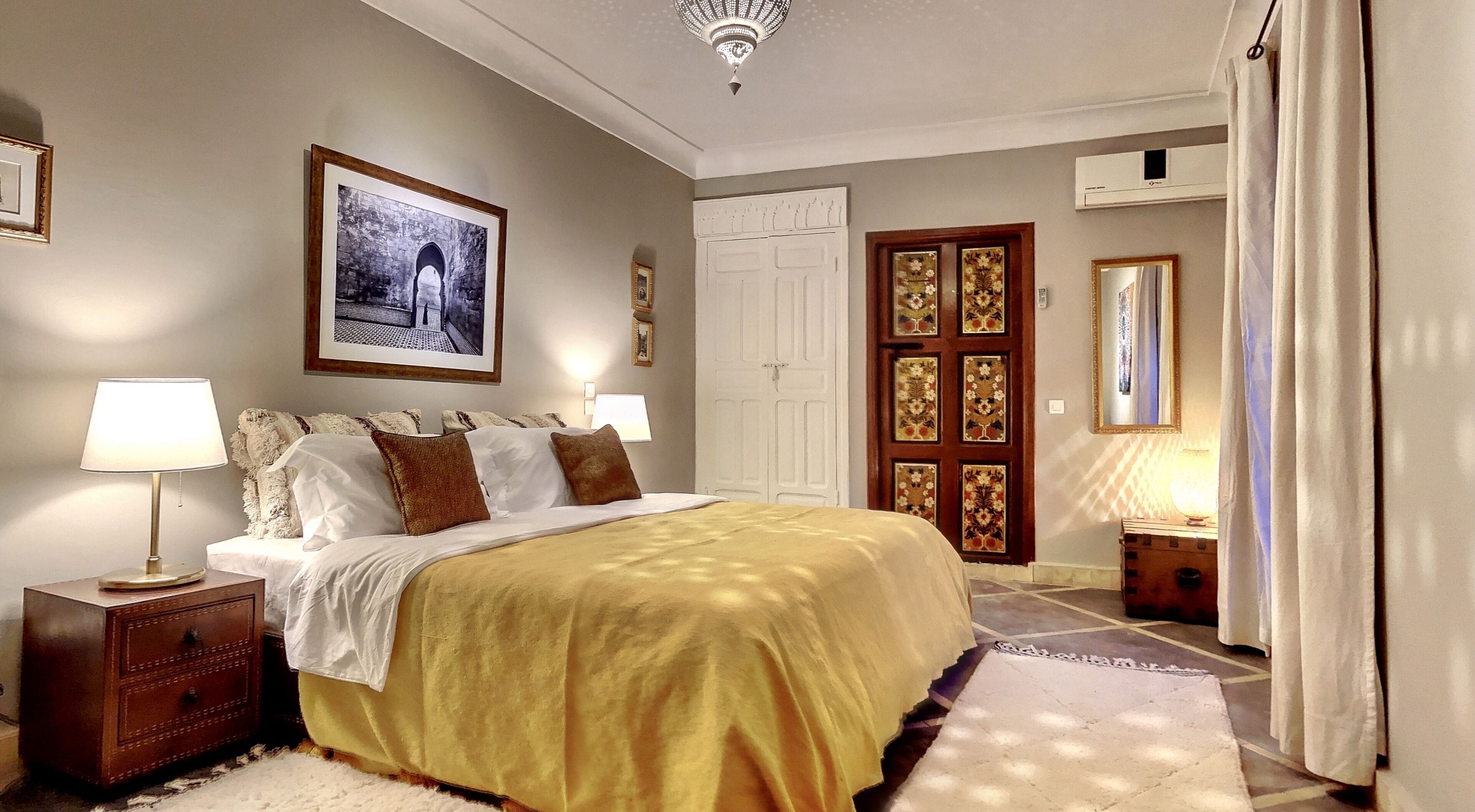 A bedroom with a bed, two bedside tables, a nightstand, a large framed picture, a door with decorative panels, and a mirror.