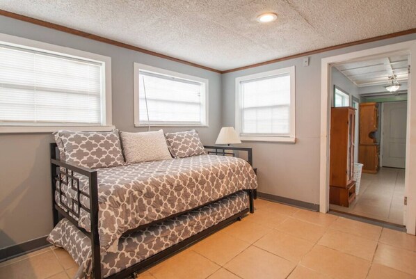 A bedroom with a bed, two windows, a lamp, and a door leading to another room.