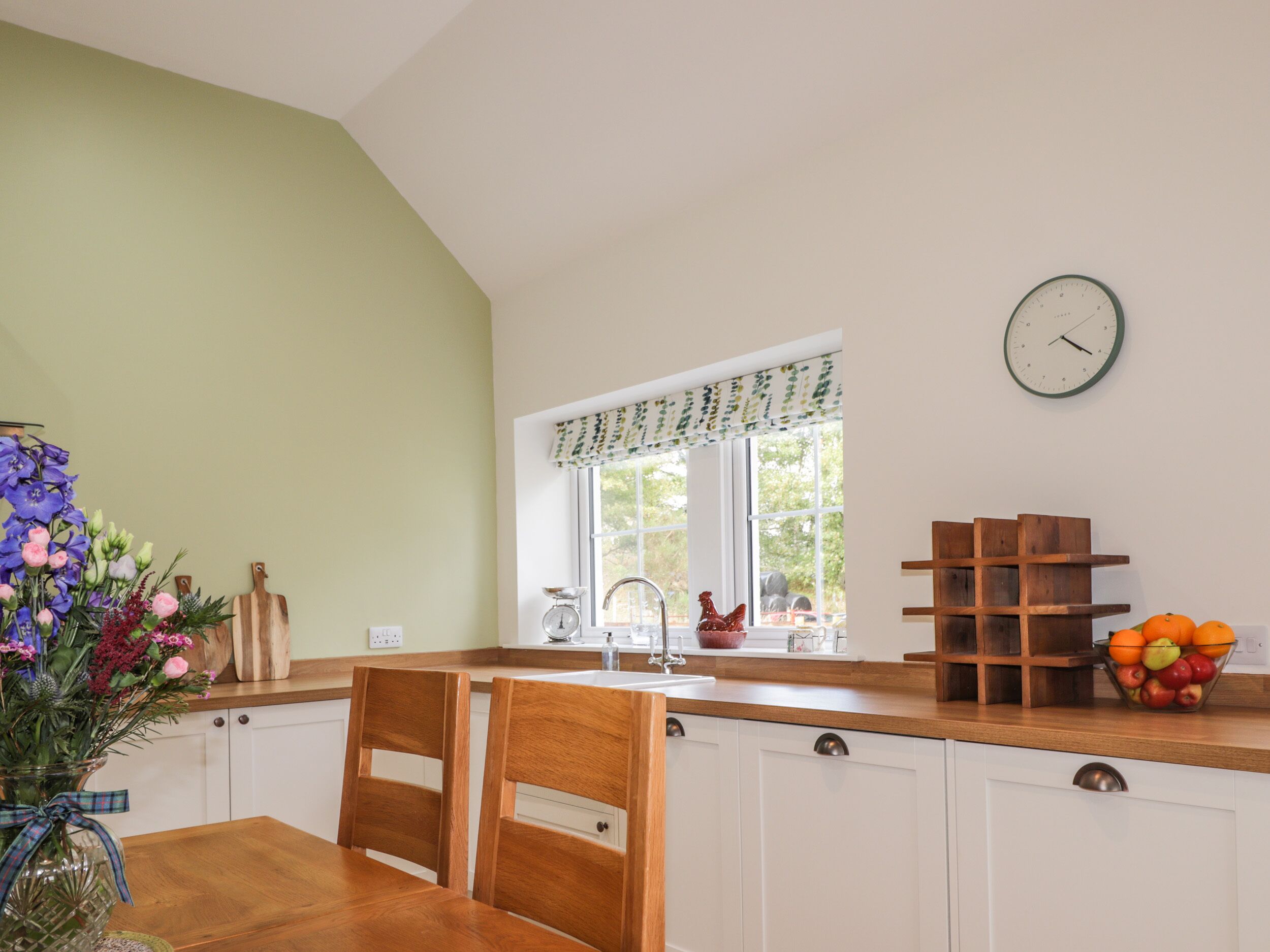 A kitchen with white cabinets, a wooden countertop, a window with a view, a clock, and a vase with flowers.