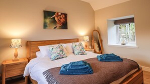 A bedroom with a wooden bed, floral pillows, a large painting, two bedside lamps, and a window with a view of trees. - Bonnach (Muir of Ord)