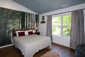 A bedroom with a large window, a bed with white bedding, a forest mural on the wall, and a small bedside table with a lamp.
