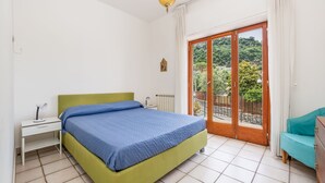 Un dormitorio con cama, mesita de noche, escritorio, silla y balcón con vistas a la vegetación.