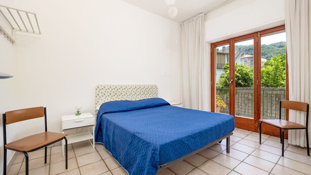 Un dormitorio con cama, dos sillas, una mesita de noche y ventana con vistas a follaje.