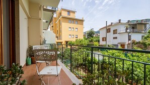 Un balcón con una silla y una mesa de metal, rodeado de follaje y vistas a edificios cercanos.