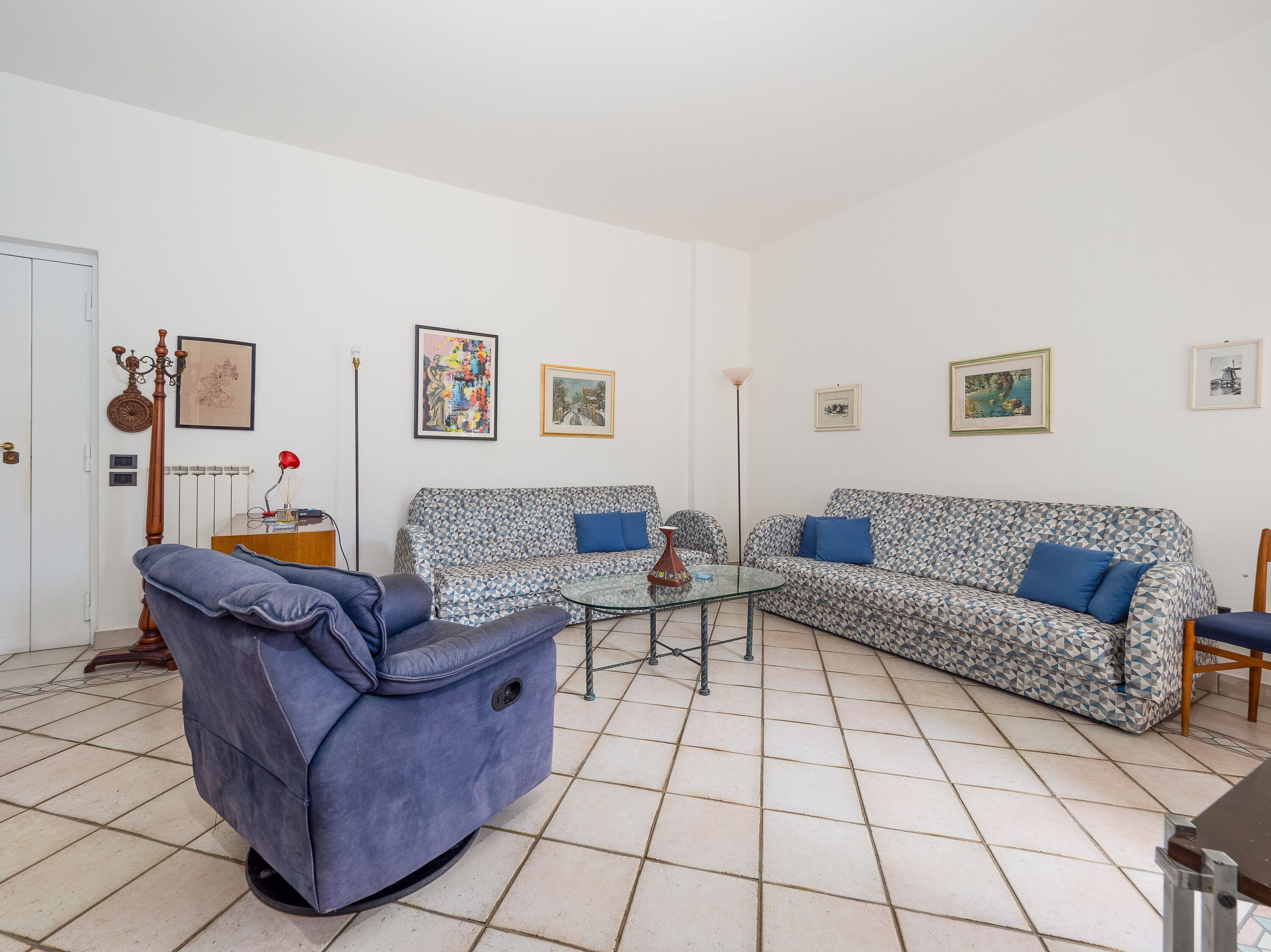 Una sala de estar con un sofá estampado, un sillón azul, una mesa de centro de vidrio y una lámpara de pie.